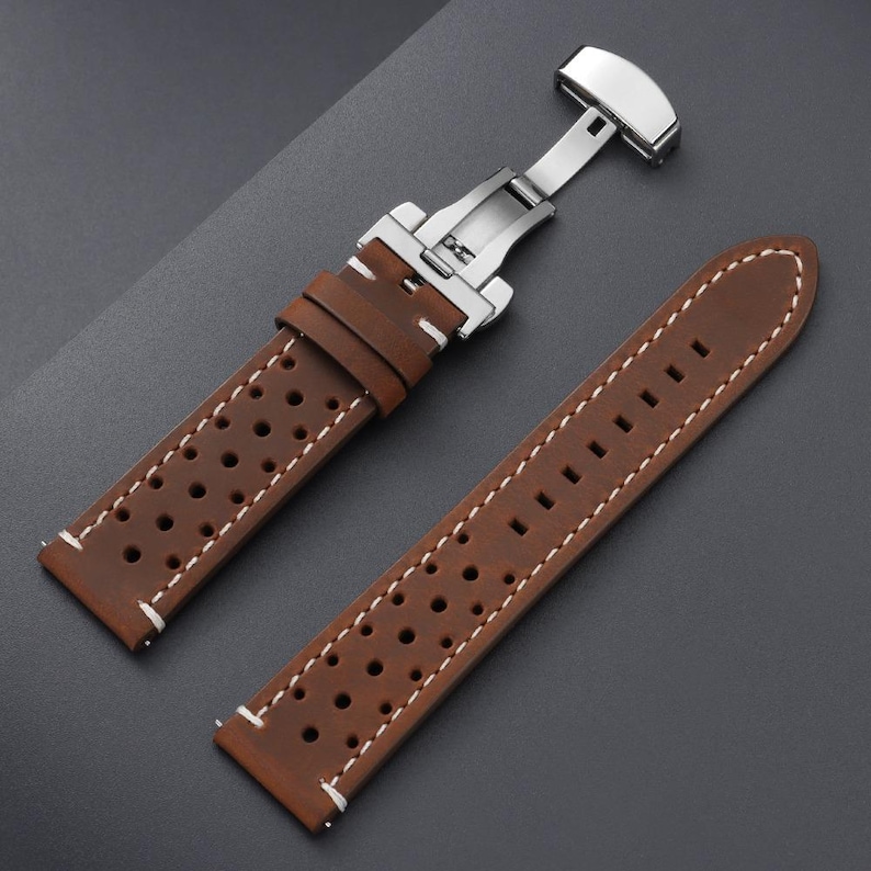 May include: Brown leather watch strap with white stitching and a stainless steel clasp. The strap features a perforated design and is shown open, displaying the clasp mechanism. The watch band is designed for a classic look.