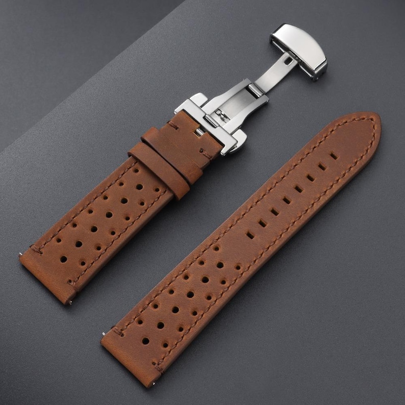 May include: A brown leather watch strap with a stainless steel butterfly clasp. The strap has a perforated design and is shown in two pieces, with the clasp open. The leather has a textured appearance.