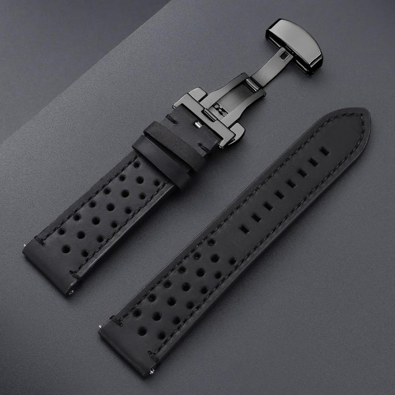 May include: Black leather watch strap with a black metal clasp. The strap has a perforated design and is shown in two pieces, with the clasp open. The background is a dark gray surface.