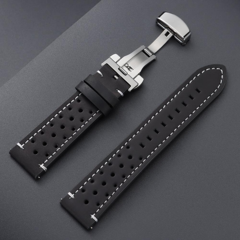 May include: Black watch strap with white stitching and a stainless steel clasp. The strap features a perforated design for breathability and a secure fit. The watch band is made of a durable material.