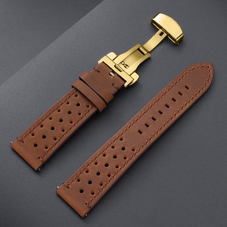 May include: Brown leather watch strap with a gold-colored clasp. The strap has a perforated design and visible stitching. The clasp is open, showing the mechanism. The watch strap is a classic accessory.
