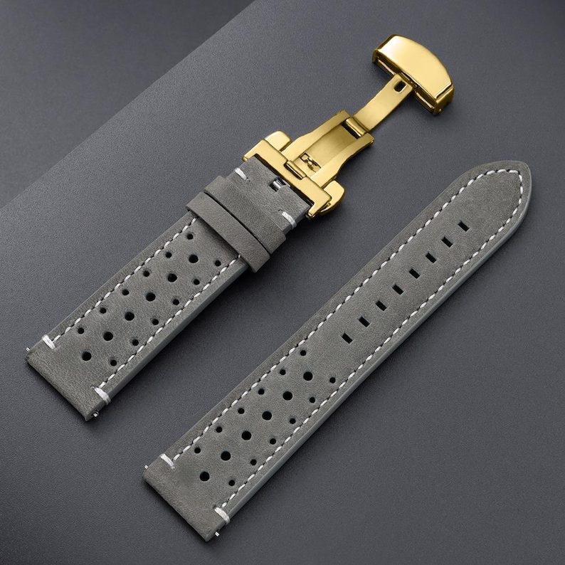 May include: Gray leather watch strap with white stitching and a gold-colored clasp. The strap has a perforated design and is made of a soft, suede-like material. The clasp is a butterfly style.