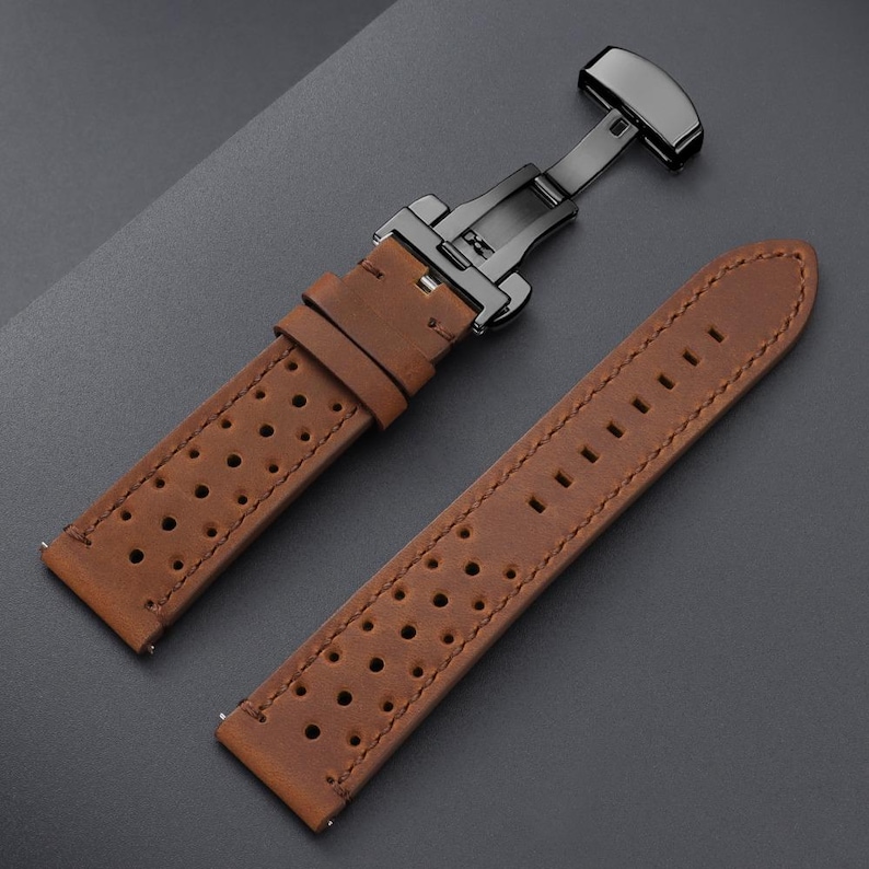 May include: Brown leather watch strap with a black clasp. The strap features a perforated design and visible stitching. The watch band is shown against a dark gray background.
