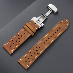 May include: A brown leather watch strap with a stainless steel clasp. The strap has a perforated design and white stitching. The watch band is shown against a dark gray background.