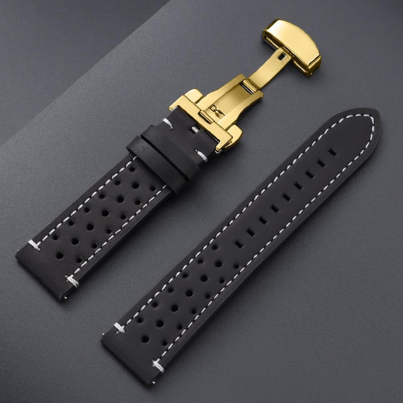 May include: Black leather watch strap with white stitching and a gold-colored clasp. The strap features a perforated design for breathability and a secure fit. The watch band is designed to be durable and stylish.