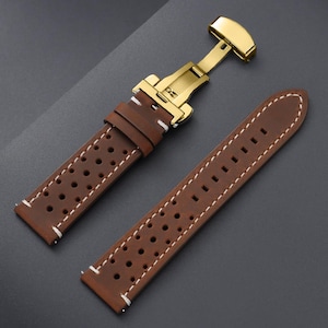 May include: Brown leather watch strap with white stitching and a gold-colored clasp. The strap features a perforated design and is shown unattached, displaying the buckle and the strap's two pieces.