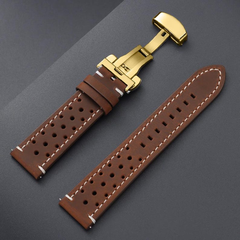 May include: Brown leather watch strap with white stitching and a gold-colored clasp. The strap features a perforated design and is shown unattached, displaying the buckle and the strap's two pieces.