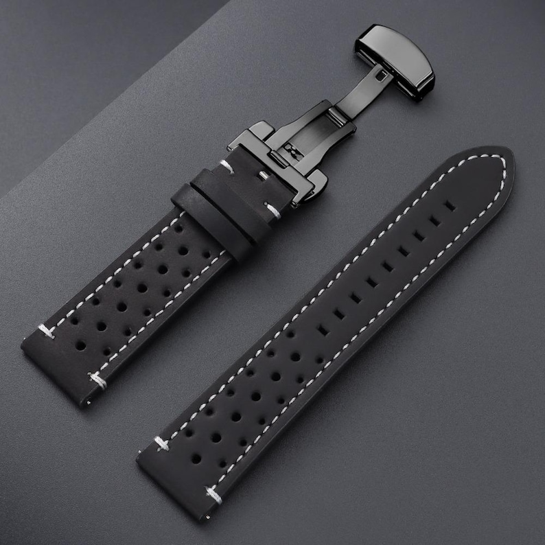 May include: Black watch strap with white stitching and a black clasp. The strap features a perforated design and is made of a smooth, matte material. The clasp is a butterfly style.