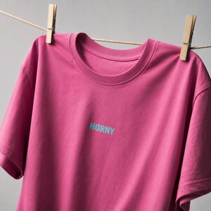 Può includere: Una maglietta magenta con la scritta "HORNY" stampata in azzurro. La maglietta è appesa a un filo con mollette di legno. Lo sfondo è grigio uniforme.