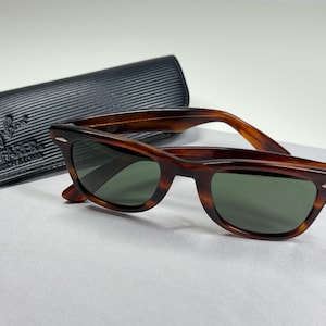 May include: Tortoiseshell-patterned Wayfarer sunglasses with dark green lenses and silver-tone accents at the hinges. The sunglasses are accompanied by a black, textured, rectangular protective case featuring the Ray-Ban Wayfarer logo embossed.