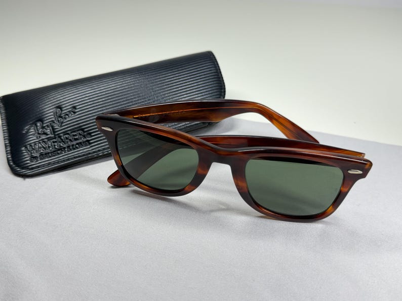 May include: Tortoiseshell-patterned Wayfarer sunglasses with dark green lenses and silver-tone accents at the hinges. The sunglasses are accompanied by a black, textured, rectangular protective case featuring the Ray-Ban Wayfarer logo embossed.