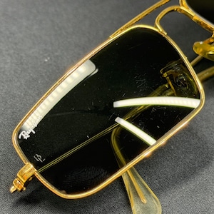 May include: Gold-tone metal sunglasses featuring a rectangular frame with a double bridge design. The dark lenses display the white Ray-Ban logo in the upper corner and a small etched symbol near the bottom. The nose pads are translucent yellow.