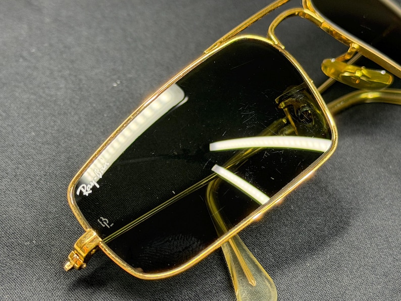 May include: Gold-tone metal sunglasses featuring a rectangular frame with a double bridge design. The dark lenses display the white Ray-Ban logo in the upper corner and a small etched symbol near the bottom. The nose pads are translucent yellow.