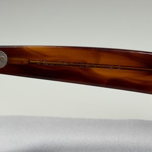 May include: A close-up view of the inner temple of brown tortoise shell patterned sunglasses. The text B&L RAY-BAN U.S.A. is engraved into the plastic arm near the silver-toned metal hinge, which features two small circular rivets.