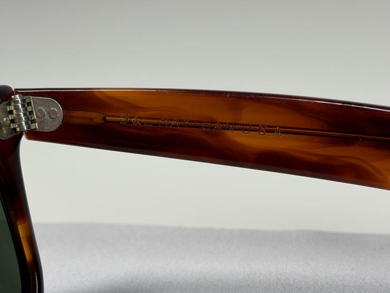 May include: A close-up view of the inner temple of brown tortoise shell patterned sunglasses. The text B&L RAY-BAN U.S.A. is engraved into the plastic arm near the silver-toned metal hinge, which features two small circular rivets.