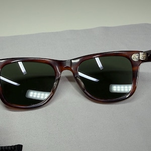 May include: Tortoiseshell-patterned sunglasses with dark green lenses and silver-toned metal hinges. The frames feature a classic rectangular shape with rounded edges and a bridge connecting the two lenses, showcasing a distinct brown and black mottled pattern throughout the frame structure.