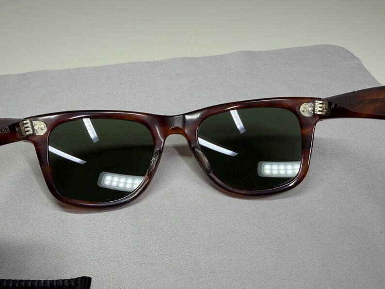May include: Tortoiseshell-patterned sunglasses with dark green lenses and silver-toned metal hinges. The frames feature a classic rectangular shape with rounded edges and a bridge connecting the two lenses, showcasing a distinct brown and black mottled pattern throughout the frame structure.
