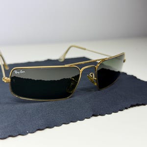 May include: Gold-tone metal frame sunglasses with a rectangular shape and dark green lenses. The top left lens features the white Ray-Ban logo. The thin wire temples and bridge connect the frames, which are resting on a dark blue fabric cloth.
