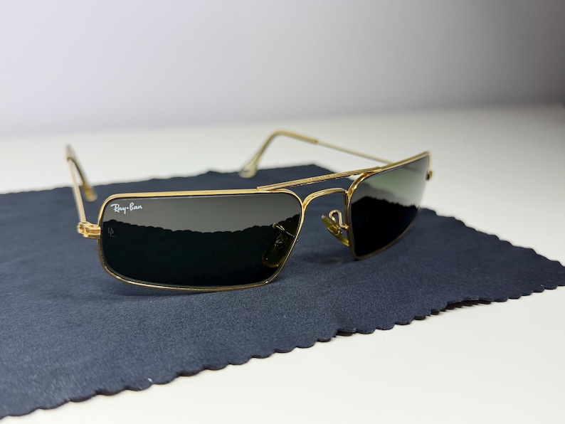 May include: Gold-tone metal frame sunglasses with a rectangular shape and dark green lenses. The top left lens features the white Ray-Ban logo. The thin wire temples and bridge connect the frames, which are resting on a dark blue fabric cloth.