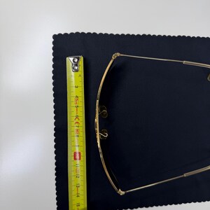May include: Gold-toned metal eyeglasses with thin frames and adjustable nose pads. A yellow measuring tape is positioned alongside the glasses, showing a length of approximately 6 inches from the top to the bottom of the frame structure.