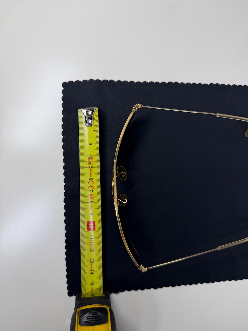 May include: Gold-toned metal eyeglasses with thin frames and adjustable nose pads. A yellow measuring tape is positioned alongside the glasses, showing a length of approximately 6 inches from the top to the bottom of the frame structure.