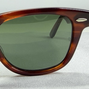 May include: Tortoiseshell-patterned sunglasses with a brown frame and green-tinted lenses. The frame features a silver-toned metal accent on the upper corner near the hinge. The lenses are dark green and show a small etched logo on the right side.