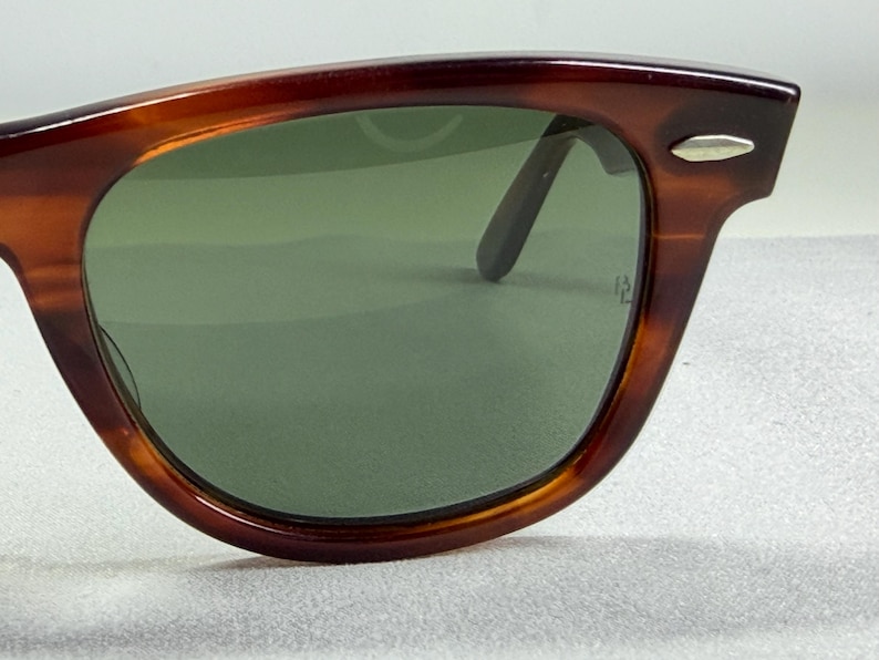 May include: Tortoiseshell-patterned sunglasses with a brown frame and green-tinted lenses. The frame features a silver-toned metal accent on the upper corner near the hinge. The lenses are dark green and show a small etched logo on the right side.