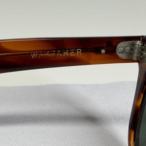 May include: Tortoiseshell-patterned sunglasses with a brown frame featuring the word WAYFARER embossed on the inner temple arm. The frame includes a silver-toned metal hinge with two visible rivets connecting the arm to the front lens section.