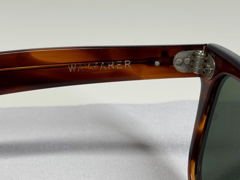 May include: Tortoiseshell-patterned sunglasses with a brown frame featuring the word WAYFARER embossed on the inner temple arm. The frame includes a silver-toned metal hinge with two visible rivets connecting the arm to the front lens section.
