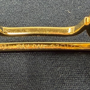 May include: Gold-tone metal bridge of eyeglasses featuring the engraved text B&L RAY-BAN 57-17. The frame includes two translucent yellow nose pads attached to the metallic structure, which is positioned horizontally against a dark textured surface.