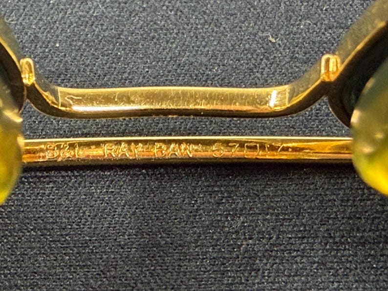 May include: Gold-tone metal bridge of eyeglasses featuring the engraved text B&L RAY-BAN 57-17. The frame includes two translucent yellow nose pads attached to the metallic structure, which is positioned horizontally against a dark textured surface.