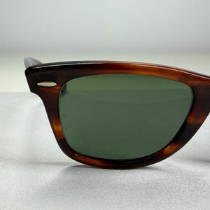 May include: Tortoiseshell-patterned sunglasses featuring a dark brown and amber frame with a glossy finish. The lenses are a solid dark green color. A small silver-toned metal accent is visible on the upper corner of the frame near the temple.