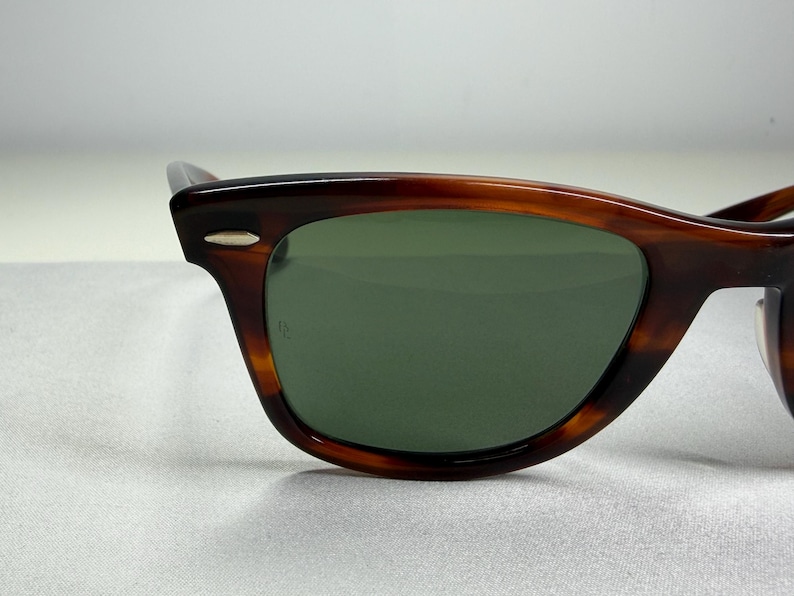 May include: Tortoiseshell-patterned sunglasses featuring a dark brown and amber frame with a glossy finish. The lenses are a solid dark green color. A small silver-toned metal accent is visible on the upper corner of the frame near the temple.
