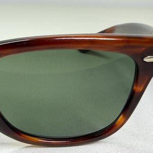 May include: Sunglasses featuring a brown tortoise shell pattern frame with a smooth, polished finish. The lenses are a solid dark green color. A small, silver-toned metallic accent is visible on the outer corner of the frame near the temple hinge.