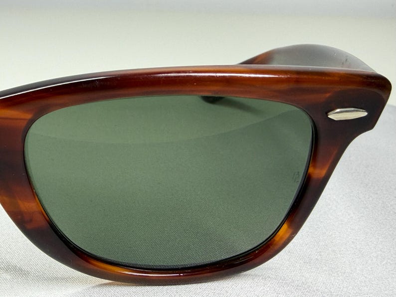 May include: Sunglasses featuring a brown tortoise shell pattern frame with a smooth, polished finish. The lenses are a solid dark green color. A small, silver-toned metallic accent is visible on the outer corner of the frame near the temple hinge.