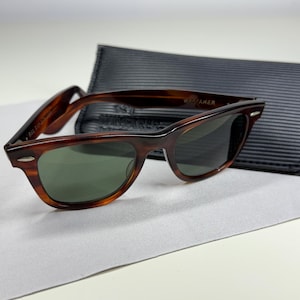 May include: Tortoiseshell-patterned sunglasses with dark green lenses and silver-tone accents at the hinges. The frames feature a classic rectangular shape with a thick bridge. A black, textured, rectangular protective case is positioned behind them.