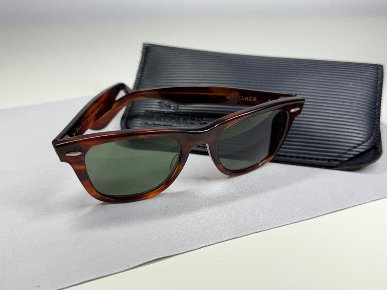 May include: Tortoiseshell-patterned sunglasses with dark green lenses and silver-tone accents at the hinges. The frames feature a classic rectangular shape with a thick bridge. A black, textured, rectangular protective case is positioned behind them.