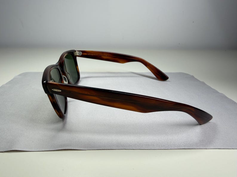 May include: Tortoiseshell-patterned sunglasses with dark green lenses and a classic frame shape. The temples feature a small silver-toned metal accent near the hinges, and the frame has a smooth, polished finish with a distinct brown and black pattern.