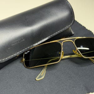 May include: Gold-tone metal sunglasses with rectangular dark lenses and a double bridge design. The left lens features the Ray-Ban logo in white. The sunglasses are accompanied by a black leather-textured hard case and a black microfiber cloth.