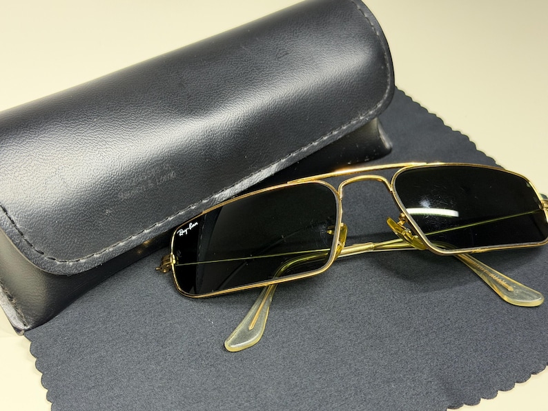 May include: Gold-tone metal sunglasses with rectangular dark lenses and a double bridge design. The left lens features the Ray-Ban logo in white. The sunglasses are accompanied by a black leather-textured hard case and a black microfiber cloth.