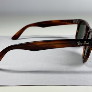 May include: Tortoiseshell sunglasses with a brown and black mottled pattern. The frames feature a classic shape with dark tinted lenses. The Ray-Ban logo is printed in silver lettering on the outer side of the temple arm near the hinge.