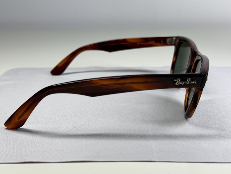 May include: Tortoiseshell sunglasses with a brown and black mottled pattern. The frames feature a classic shape with dark tinted lenses. The Ray-Ban logo is printed in silver lettering on the outer side of the temple arm near the hinge.