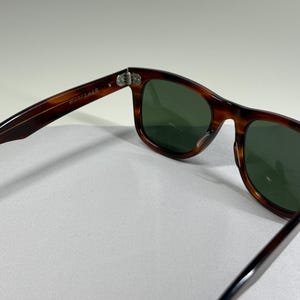 May include: Tortoiseshell-patterned sunglasses with dark green lenses. The inner temple arm features the text "WAYFARER" printed in white. The frame has a classic rectangular shape with silver-toned metal hinges connecting the arms to the front.