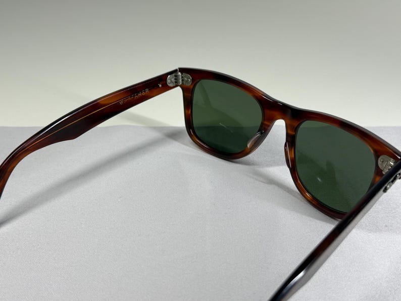 May include: Tortoiseshell-patterned sunglasses with dark green lenses. The inner temple arm features the text "WAYFARER" printed in white. The frame has a classic rectangular shape with silver-toned metal hinges connecting the arms to the front.