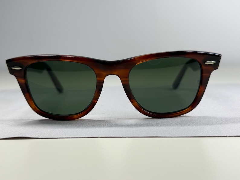 May include: Tortoiseshell-patterned sunglasses with a brown and black frame and dark green lenses. The frame features a classic wayfarer shape with silver-toned metal accents on the outer corners of the front rims, providing a distinct look.