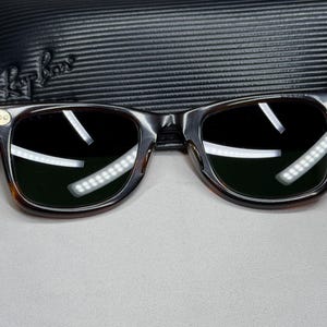May include: Dark brown tortoise shell sunglasses with dark green lenses and gold-tone metal accents at the hinges. The frames feature a classic rectangular shape with rounded edges. A black textured Ray-Ban branded carrying case is positioned behind.