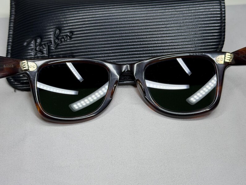 May include: Dark brown tortoise shell sunglasses with dark green lenses and gold-tone metal accents at the hinges. The frames feature a classic rectangular shape with rounded edges. A black textured Ray-Ban branded carrying case is positioned behind.