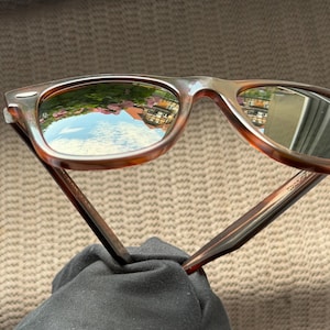 May include: Tortoiseshell-patterned sunglasses with dark-tinted lenses. The lenses feature a reflective surface showing a clear blue sky, green foliage, and a building. The frames have a classic rectangular shape with silver-toned metal accents.