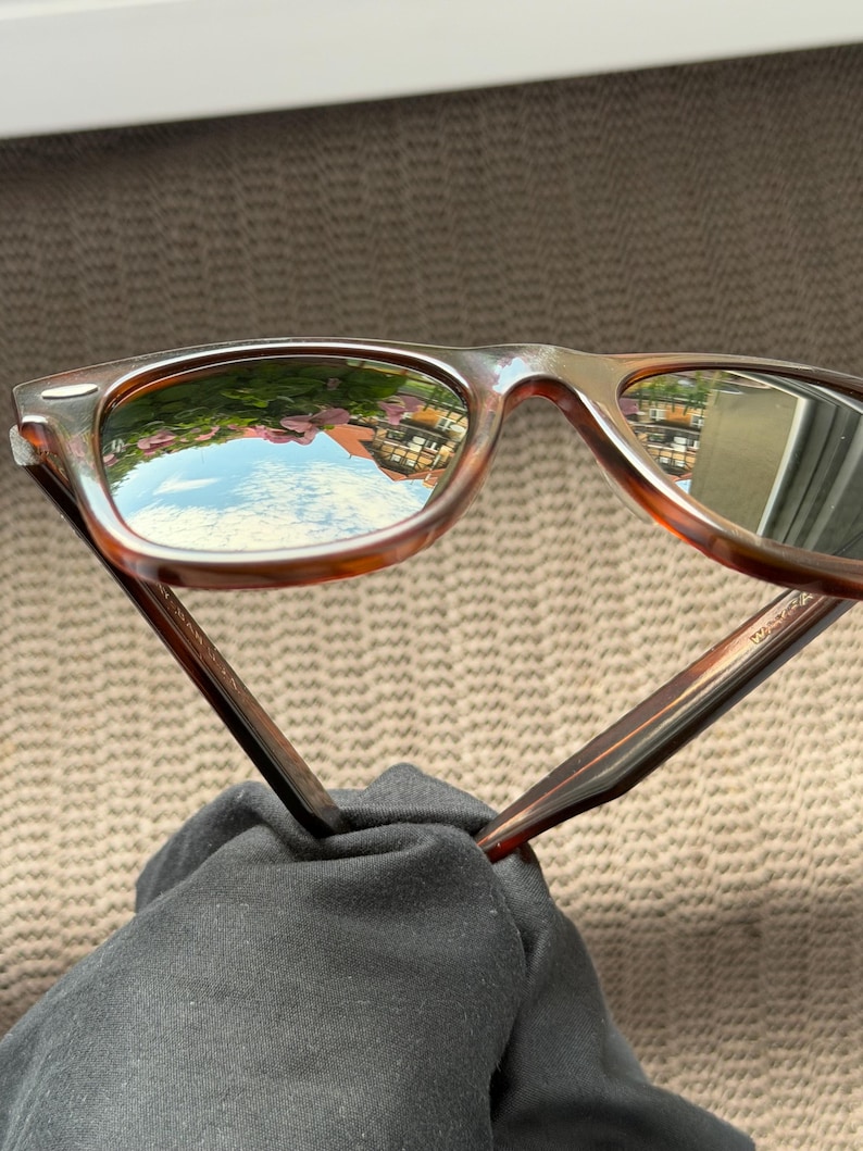 May include: Tortoiseshell-patterned sunglasses with dark-tinted lenses. The lenses feature a reflective surface showing a clear blue sky, green foliage, and a building. The frames have a classic rectangular shape with silver-toned metal accents.