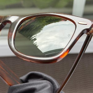 May include: Tortoiseshell-patterned sunglasses with dark green lenses. The frame features a classic rectangular shape with a small silver-toned metal accent on the top corner of the lens rim. The lenses show a reflection of the outdoors.